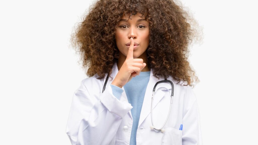 Una doctora con bata blanca y estetoscopio haciendo el gesto de silencio con el dedo índice sobre los labios.