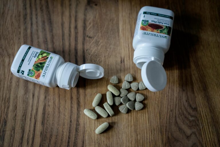 Dos botes blancos de suplementos Nutrilite, "Daily" e "Iron Folic Plus", sobre una mesa de madera con varias pastillas verdes y marrones esparcidas entre ellos.
