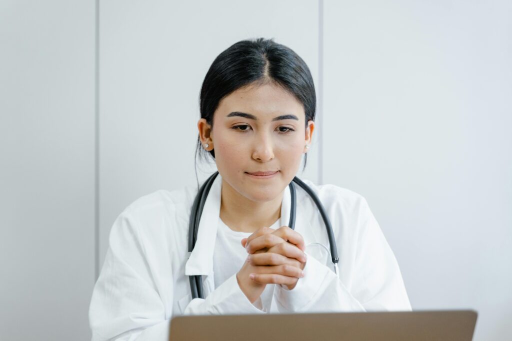 Una profesional de la salud con bata blanca y estetoscopio sentada frente a un ordenador portátil, representando la telemedicina y consultas de salud.