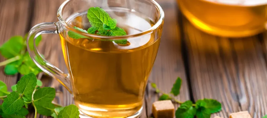 Una taza de vidrio transparente con té de color ámbar, decorada con hojas de ortiga fresca y una flor amarilla sobre un posavasos de arpillera.