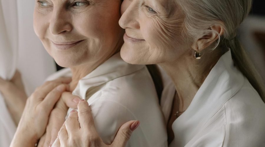 Primer plano de dos mujeres mayores sonrientes y abrazadas, vistiendo camisas blancas, representando el apoyo familiar y la salud emocional.