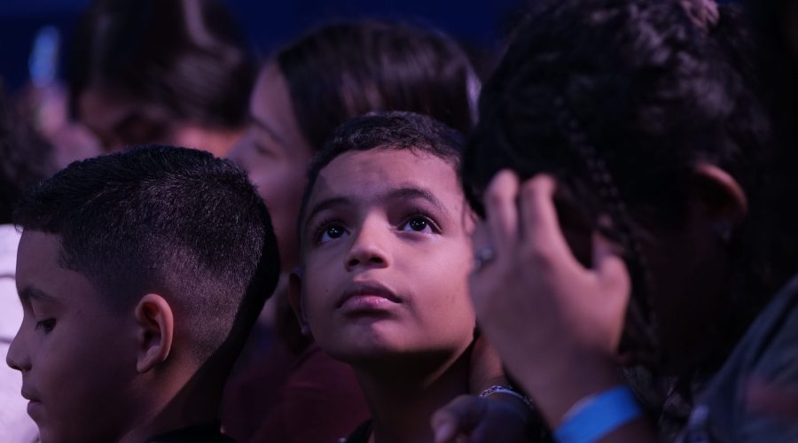 "Primer plano de un niño de tez morena mirando hacia arriba con expresión de asombro y atención, rodeado de otras personas en un entorno con iluminación tenue, preparándose para un evento como un eclipse