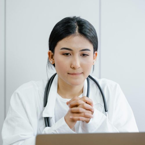 Una profesional de la salud con bata blanca y estetoscopio sentada frente a un ordenador portátil, representando la telemedicina y consultas de salud.